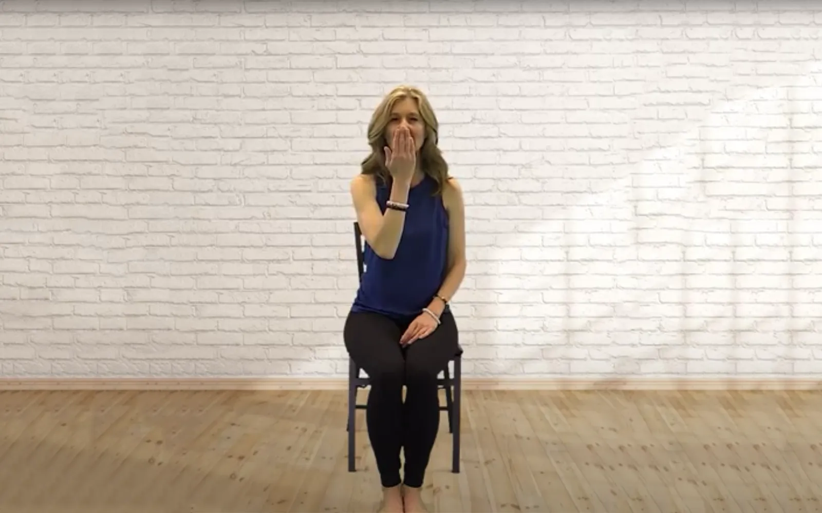 A woman sits on a chair with her legs relaxed, one hand on her lap and one in front of her mouth as she inhales.